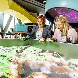 zwei Frauen stehen mit Lupen in der Hand vor einer Art Sandkiste. In der Sandkiste kann eine Landschaft geformt werden. Es können Lupen über die Oberfläche gehalten werden, die verschiedene Effekte auslösen. Jedoch wird nur ein Bild projiziert.