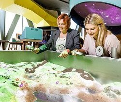 zwei Frauen stehen mit Lupen in der Hand vor einer Art Sandkiste. In der Sandkiste kann eine Landschaft geformt werden. Es können Lupen über die Oberfläche gehalten werden, die verschiedene Effekte auslösen. Jedoch wird nur ein Bild projiziert.