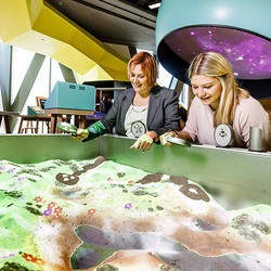 zwei Frauen stehen mit Lupen in der Hand vor einer Art Sandkiste. In der Sandkiste kann eine Landschaft geformt werden. Es können Lupen über die Oberfläche gehalten werden, die verschiedene Effekte auslösen. Jedoch wird nur ein Bild projiziert.