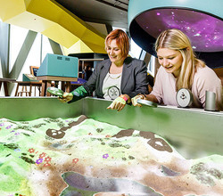 zwei Frauen stehen mit Lupen in der Hand vor einer Art Sandkiste. In der Sandkiste kann eine Landschaft geformt werden. Es können Lupen über die Oberfläche gehalten werden, die verschiedene Effekte auslösen. Jedoch wird nur ein Bild projiziert.