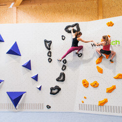 Zwei Frauen in Sportkleidung klettern an einer Indoor Wand mit verschiedenen Schwierigkeitsgraden. Die Wand ist weiß die Kletterelemente sind in blau, schwarz und orange gehalten