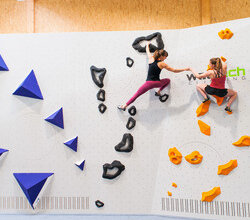 Zwei Frauen in Sportkleidung klettern an einer Indoor Wand mit verschiedenen Schwierigkeitsgraden. Die Wand ist weiß die Kletterelemente sind in blau, schwarz und orange gehalten