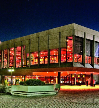 Das Bild zeigt das Theatergebäude von Heilbronn in der Nacht. Große Glasfassaden lassen das warme Licht aus dem Inneren nach außen strahlen. Der umgebende Platz ist gepflastert, und der dunkle Nachthimmel hebt das beleuchtete Gebäude hervor.