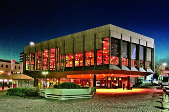 Das Bild zeigt das Theatergebäude von Heilbronn in der Nacht. Große Glasfassaden lassen das warme Licht aus dem Inneren nach außen strahlen. Der umgebende Platz ist gepflastert, und der dunkle Nachthimmel hebt das beleuchtete Gebäude hervor.