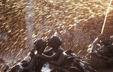 Das Bild zeigt eine detailreiche Brunnenfigur, die zwei Personen darstellt, die sich umarmen. Goldene Wassertropfen sprühen im Hintergrund in einem funkelnden Lichtbogen, was der Szene eine lebendige und magische Atmosphäre verleiht.