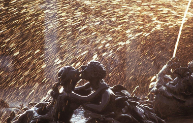 Das Bild zeigt eine detailreiche Brunnenfigur, die zwei Personen darstellt, die sich umarmen. Goldene Wassertropfen sprühen im Hintergrund in einem funkelnden Lichtbogen, was der Szene eine lebendige und magische Atmosphäre verleiht.