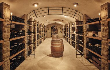 eleganter Gewölbekeller mit mehreren, gut bestückten, Weinregalen rechts und links. In der Mitte des Kellers steht ein Weinfass | © Weingut Albrecht-Kiessling