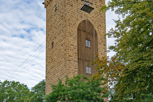 Im Fokus steht ein hoher Turm aus Naturstein. Auf einer Seite sind die Steine durch eine bogenförmigen Holzkonstruktion mit Fenster unterbrochen. Auf einer weiteren Seite ist eine Uhr zu sehen. Vor dem Turm sind auf dem Gehweg Bäume gepflanzt