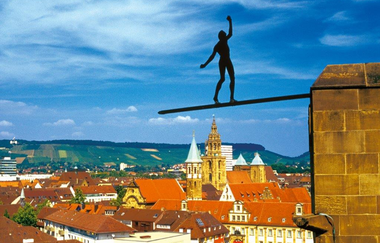 Das Bild zeigt die scherenschnittartige Silhouette einer Person, die auf einem schmalen Balken balanciert. Der Balken ist an einer Mauer befestigt. Im Hintergrund sind Teile der Stadt, vorwiegend die Dächer sichtbar. In der Ferne sieht man die Weinberge