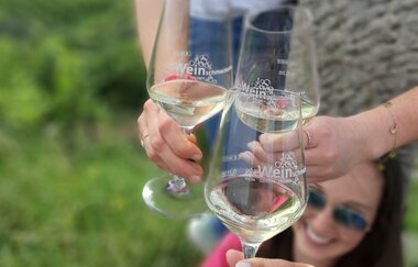 Im Focus stehen 3 Sektgläser, mit denen angestoßen wird. Im Hintergrund erkennt man unscharf eine junge Frau mit Sonnenbrille, die entspannt lächelt. | © DieWeinschmeckerin Nicole Halter