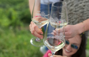 Im Focus stehen 3 Sektgläser, mit denen angestoßen wird. Im Hintergrund erkennt man unscharf eine junge Frau mit Sonnenbrille, die entspannt lächelt. | © DieWeinschmeckerin Nicole Halter