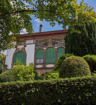 Die Villa Schliz ist im floralen Jugendstil erbaut. Auf dem Bild sind die bogenartigen Fenster mit grünen Rollladen zu erkennen. Die Villa liegt geschützt hinter einer Hecke und verschiedenen in Form geschnittener Bäume