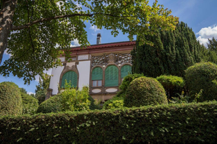 Die Villa Schliz ist im floralen Jugendstil erbaut. Auf dem Bild sind die bogenartigen Fenster mit grünen Rollladen zu erkennen. Die Villa liegt geschützt hinter einer Hecke und verschiedenen in Form geschnittener Bäume