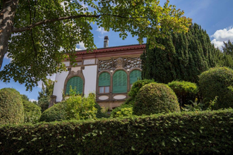 Die Villa Schliz ist im floralen Jugendstil erbaut. Auf dem Bild sind die bogenartigen Fenster mit grünen Rollladen zu erkennen. Die Villa liegt geschützt hinter einer Hecke und verschiedenen in Form geschnittener Bäume