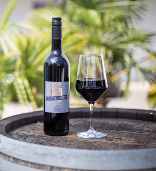 Das Bild zeigt eine Rotweinflasche und ein Glas mit Rotwein auf einem Holzfass. Im Hintergrund sind kleine Palmen zu sehen. | © Heilbronn Marketing GmbH