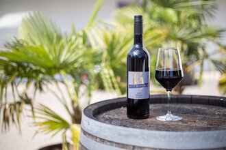 Das Bild zeigt eine Rotweinflasche und ein Glas mit Rotwein auf einem Holzfass. Im Hintergrund sind kleine Palmen zu sehen. | © Heilbronn Marketing GmbH
