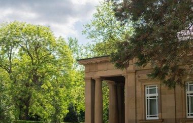 Ein Gebäude aus beigefarbenem Stein mit Säulen steht inmitten eines weitläufigen Parks unter einem bewölkten Himmel.