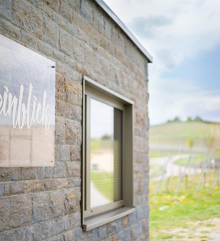 Das Bild zeigt eine Steinmauer, an der ein transparentes Schild mit der goldenen Aufschrift "Weinblick" angebracht ist. Neben dem Schild ist ein Fenster in der Mauer. Im Hintergrund sieht man die grünen Felder eines Weinbergs.