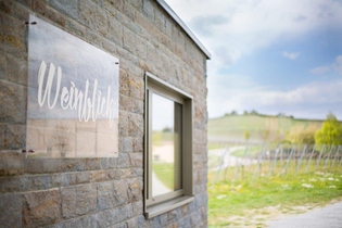 Das Bild zeigt eine Steinmauer, an der ein transparentes Schild mit der goldenen Aufschrift "Weinblick" angebracht ist. Neben dem Schild ist ein Fenster in der Mauer. Im Hintergrund sieht man die grünen Felder eines Weinbergs.