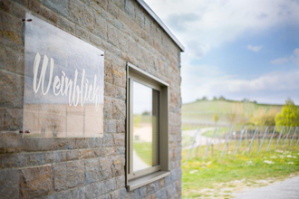 Das Bild zeigt eine Steinmauer, an der ein transparentes Schild mit der goldenen Aufschrift "Weinblick" angebracht ist. Neben dem Schild ist ein Fenster in der Mauer. Im Hintergrund sieht man die grünen Felder eines Weinbergs.