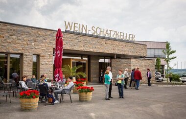 Einladende Szene vor einem Gebäude. Gäste sitzen entspannt an Tischen, umgeben von blühenden Blumenkübeln. Eine Gruppe steht am Eingang. Die Fassade ist aus Naturstein und auf dem Dach steht die Aufschrift "Wein-Schatzkeller" in goldenen Großbuchstaben.