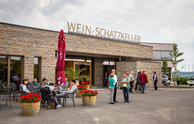 Einladende Szene vor einem Gebäude. Gäste sitzen entspannt an Tischen, umgeben von blühenden Blumenkübeln. Eine Gruppe steht am Eingang. Die Fassade ist aus Naturstein und auf dem Dach steht die Aufschrift "Wein-Schatzkeller" in goldenen Großbuchstaben.
