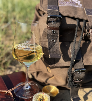 Weißwein im Glas auf einem Buch, neben dran ein Kompass und dahinter ein brauner Rucksack | © Nicole Halter