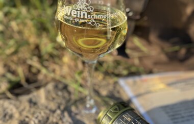 auf einem Stein steht ein Weinglas mit Weißwein, Kompass und ein Cryptex. Im Hintergrund ist ein brauner Rucksack zu sehen. | © Nicole Halter