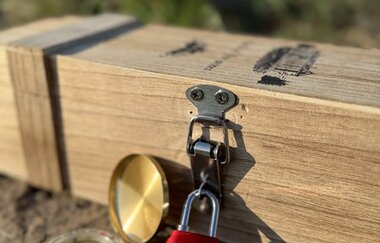 auf einem Stein liegt eine Weinkiste verschlossen mit einem roten Zahlenschloss, davor liegt noch ein Kompass | © Nicole Halter