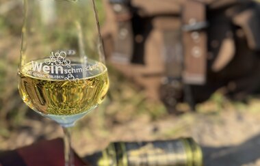 im Vordergrund ist ein Weinglas mit Weißwein zu sehen, dass auf einem braunen Buch steht, daneben ein Kompass und dahinter Cyptrex und ein brauner Rucksack | © Nicole Halter