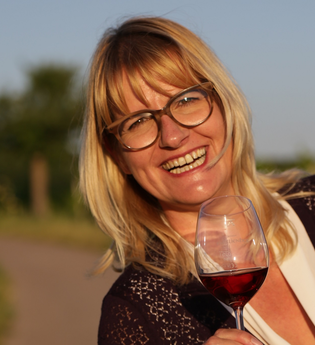 Portrait von Nicole Halter mit einem Glas Rotwein in der Hand. Mit einem gewinnenden Lächeln strahlt sie mit der Sonne um die Wette. | © DieWeinschmeckerin Nicole Halter