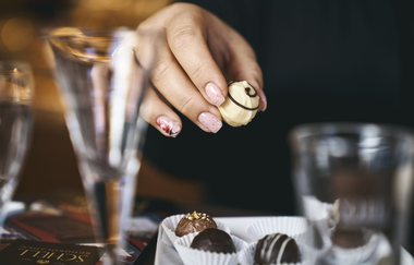 Im Fokus des Imagebildes ist eine Hand, die eine weiße Praline hält. Im Vordergrund mit Unschärfe einige Weingläser | © Tourimia Tourismus GmbH