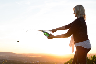 Nicole Halter sabriert eine Champagnerflasche bei Sonnenuntergang auf einem Weinberg. Der fliegende Korken und das spritzende Getränk betonen die Dynamik. Die warme Beleuchtung und die weite Landschaft schaffen eine feierliche, elegante Stimmung. | © DieWeinschmeckerin Nicole Halter