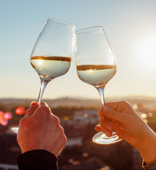 Im Fokus des Bildes sind die Hände von unterschiedlichen Personen zu sehen, die jeweils ein Weinglas mit Weißwein halten  und miteinander anstoßen. Verschwommen im Hintergrund erahnt man die Stadt in der untergehenden Sonne.