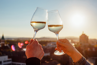 Im Fokus des Bildes sind die Hände von unterschiedlichen Personen zu sehen, die jeweils ein Weinglas mit Weißwein halten  und miteinander anstoßen. Verschwommen im Hintergrund erahnt man die Stadt in der untergehenden Sonne.