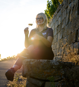 Nicole Halter sitzt entspannt auf einer Steinmauer am Rand eines Weinbergs und hält ein Glas Wein. Im Hintergrund scheint die Sonne warm durch das Grün, die Szene wirkt friedlich und idyllisch. Der Weg neben ihr führt leicht bergauf. | © Die Weinschmeckerin Nicole Halter