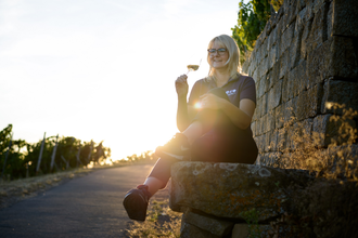Nicole Halter sitzt entspannt auf einer Steinmauer am Rand eines Weinbergs und hält ein Glas Wein. Im Hintergrund scheint die Sonne warm durch das Grün, die Szene wirkt friedlich und idyllisch. Der Weg neben ihr führt leicht bergauf. | © Die Weinschmeckerin Nicole Halter