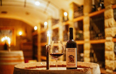 Eine Weinflasche steht neben einem Glas mit Rotwein und einer Kerze auf einem Holzfass. Im Hintergrund kann man die Regale aus Stein und Holz eines Weinkellers sehen. Entlang der Regale und auf anderen Holzfässern im Hintergrund brennen weitere Kerzen.