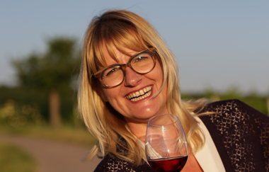 Portrait von Nicole Halter mit einem Glas Rotwein in der Hand. Mit einem gewinnenden Lächeln strahlt sie mit der Sonne um die Wette. | © DieWeinschmeckerin Nicole Halter