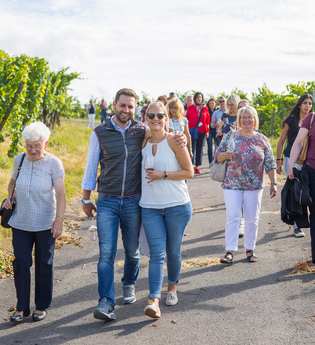 Eine Gruppe fröhlicher Menschen wandert über einen asphaltierten Weg zwischen Weinreben | © Heilbronn Marketing GmbH