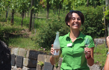 lachende Frau mit kurzen, dunklen Haaren in einem grünen Shirt. Stehend in den Weinbergen und Weinfalsche in der Hand. | © Anke Schäffer