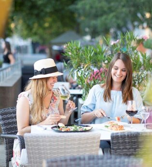 zwei Frauen sitzen nebeneinander an einem Tisch, der mit Speisen und Wein gedeckt ist. Eine Frau trägt einen beigen Sonnenhut mit dunklem Band. Links ist der Sitzplatz durch ein Geländer abgegrenzt . Hinter den beiden  stehen Blumenkübel mit Oleander
