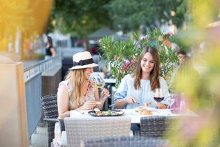zwei Frauen sitzen nebeneinander an einem Tisch, der mit Speisen und Wein gedeckt ist. Eine Frau trägt einen beigen Sonnenhut mit dunklem Band. Links ist der Sitzplatz durch ein Geländer abgegrenzt . Hinter den beiden  stehen Blumenkübel mit Oleander