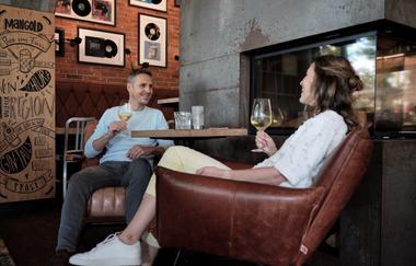 Das Bild zeigt, in seitlicher Perspektive, eine Frau mit einem Weinglas in der Hand  die in einem braunem Ledersessel sitzt. Ihr gegenüber sitzt ein Mann der auch ein Weinglas in der Hand hält. An der Holzwand dahinter hängen Kollagen mit Schallplatten