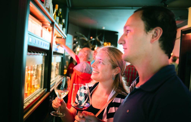 Ein Mann und eine Frau mit einem Weinglas in der Hand stehen vor einer beleuchteten, verglasten Wand mit vielen kleinen Zapfhähnen aus denen selbstständig Wein gezapft werden kann. Darüber sind Weinflaschen aufgereiht.