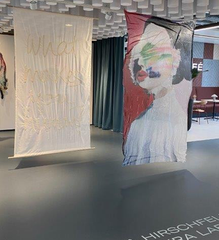Das Bild zeigt zwei große, textile Kunstwerke, die an der Decke hängen.  Eines ist weiß und mit einer Stickerei verziert, das andere zeigt ein abstraktes Bild einer Person auf einem rot-braunen Hintergrund. An der Decke hängen zylindrische Elemente.