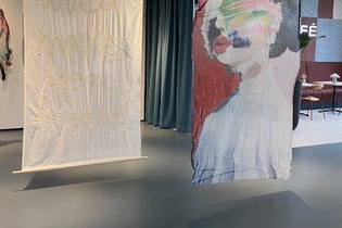 Das Bild zeigt zwei große, textile Kunstwerke, die an der Decke hängen.  Eines ist weiß und mit einer Stickerei verziert, das andere zeigt ein abstraktes Bild einer Person auf einem rot-braunen Hintergrund. An der Decke hängen zylindrische Elemente.