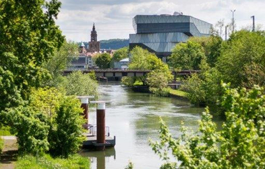 Auf dem Bild ist ein Fluss mit üppigem Grün an den Ufern zu sehen.  Im Hintergrund ragt ein modernes Gebäude (der Science Dome) heraus. Dahinter ist auch die Spitze der Kilianskirche zu sehen. Ein kleines Anlegedeck ist auf dem Wasser zu erkennen.