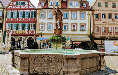 Der Milchlingsbrunnen in Bad Mergentheim. In der Mitte steht eine Statue mit einem Ritter obendrauf. Im Hintergrund sind die Fachwerkhäuser des Markplatzes zu sehen. | © Bad Mergentheim