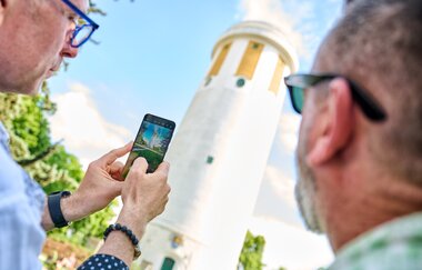 zwei Männer betrachten ein Handy mit einem Foto vom Hockenheimer Wasserturm. | © Tourimia Tourismus Gmb